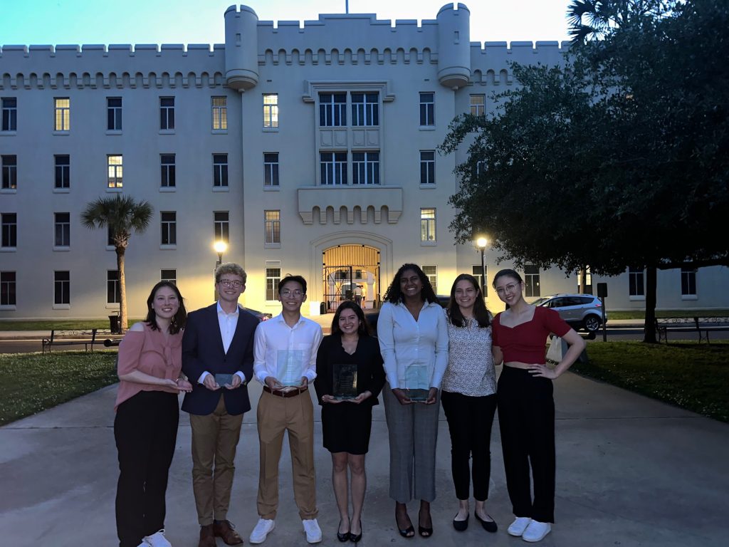 Seven students holding awards stand before the gateway of The Citidel in South Carolina at dusk. 