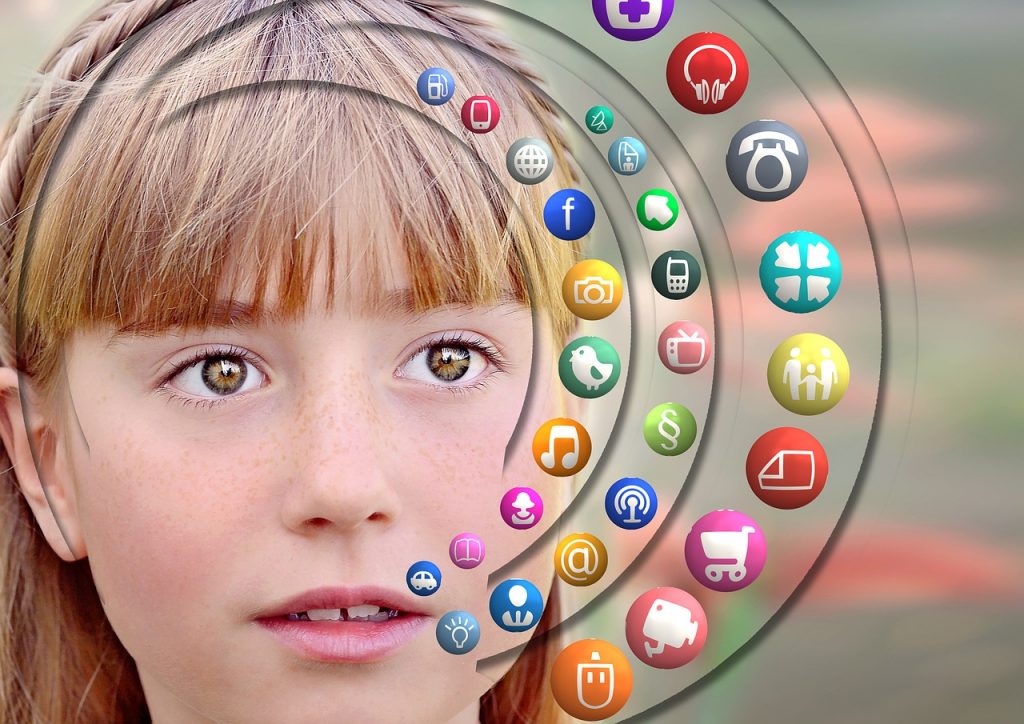 The face of a brown-eyed girl with freckles, bangs and new adult teeth fills most of the frame. Superimposed to the right are the icons of multiple real and imagined social media apps in a semicircular arrangement. Image by geralt, via Pixabay.